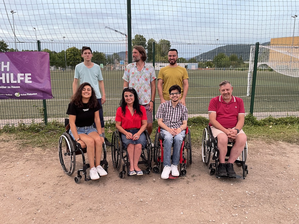 zwei Rollstuhlfahrerinnen, zwei Rollstuhlfahrer und drei Fußgänger stehen auf einem Sportplatz